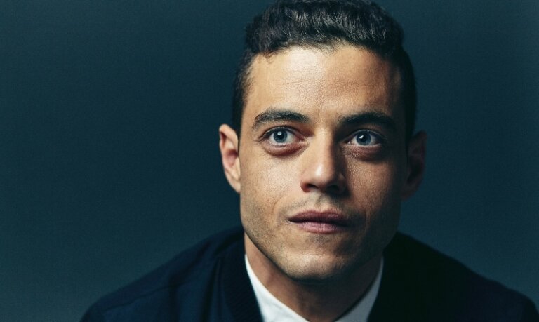 Portrait of Rami Malek in a dark jacket against a deep teal background, close-up shot highlighting his facial features and confident gaze.