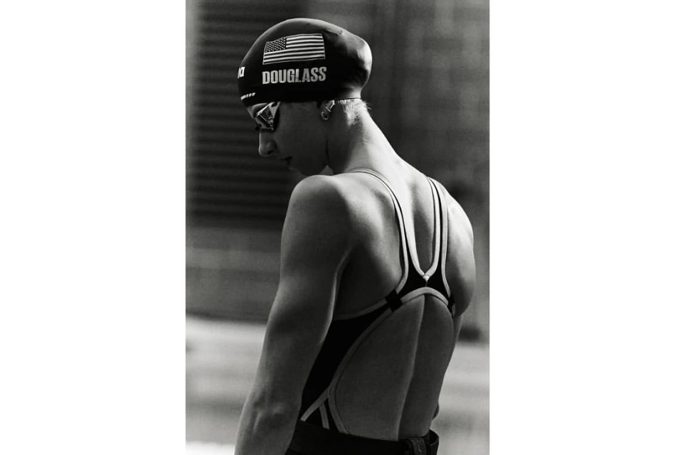 Black and white portrait of a young adult female swimmer in a swimsuit and swim cap with goggles, captured in a medium shot.