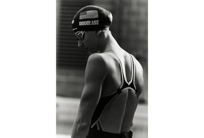 Black and white portrait of a young adult female swimmer in a swimsuit and swim cap with goggles, captured in a medium shot.