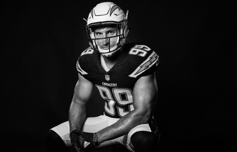 Black-and-white portrait of a football player in uniform and helmet, kneeling on one knee against a dark backdrop in a photo studio.