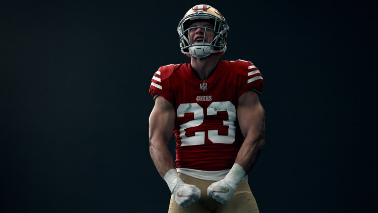 Football player in 49ers uniform flexing, showing biceps. He wears a helmet and white wristbands against a dark gray background.