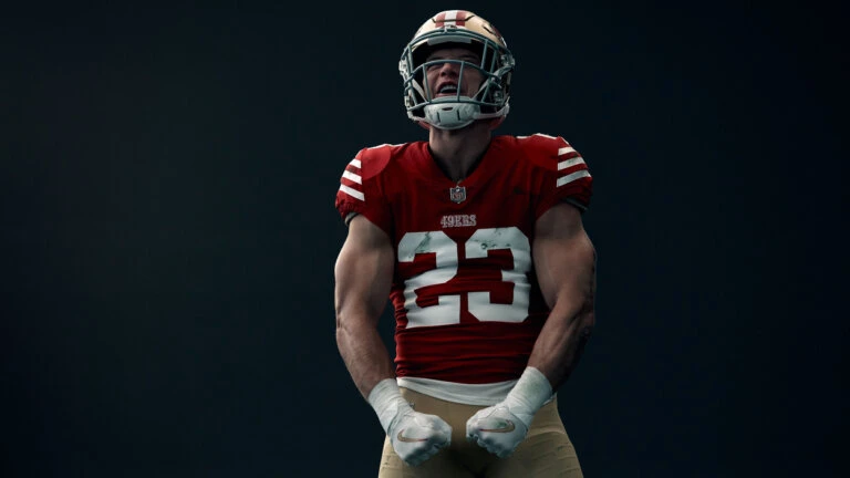 Football player in 49ers uniform flexing, showing biceps. He wears a helmet and white wristbands against a dark gray background.