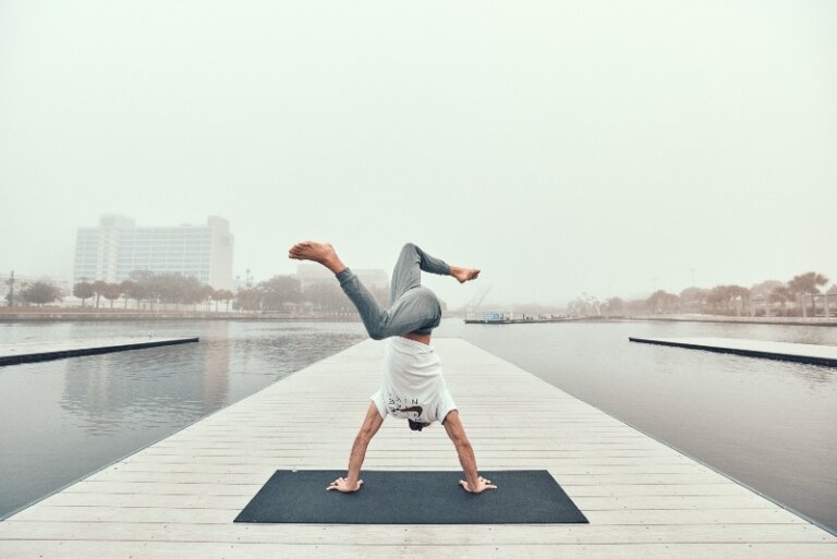An adult does a yoga handstand pose on a mat on a dock with water and a city in the background during a foggy day.