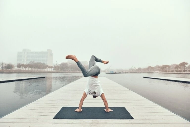 An adult does a yoga handstand pose on a mat on a dock with water and a city in the background during a foggy day.