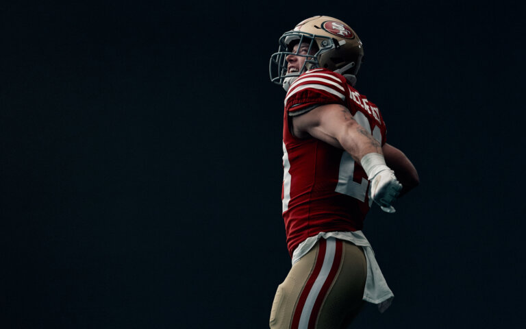 Close-up of an adult male athlete in uniform and helmet, flexing outdoors against a dark backdrop with natural light.