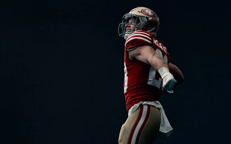 Close-up of an adult male athlete in uniform and helmet, flexing outdoors against a dark backdrop with natural light.