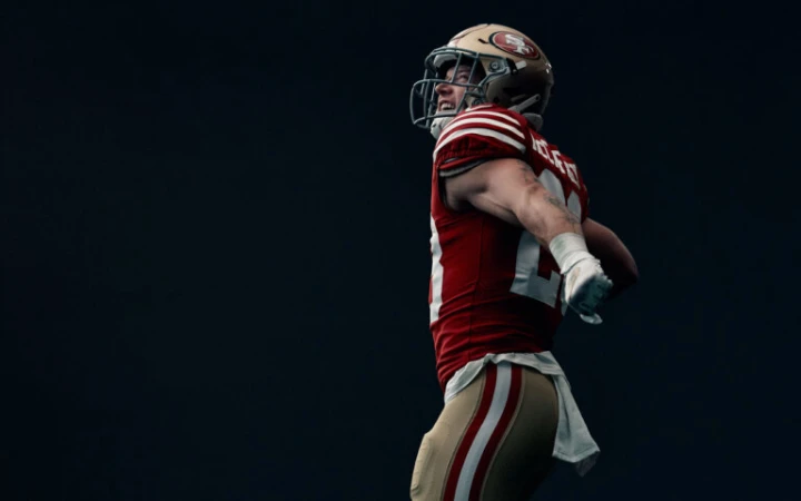 Close-up of an adult male athlete in uniform and helmet, flexing outdoors against a dark backdrop with natural light.
