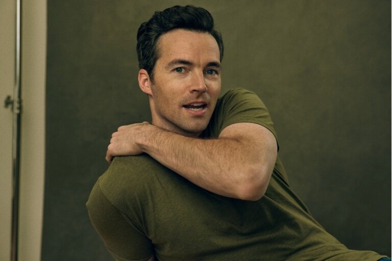 Portrait of a casually dressed adult male with dark hair, looking towards the camera against a muted olive-green studio backdrop.