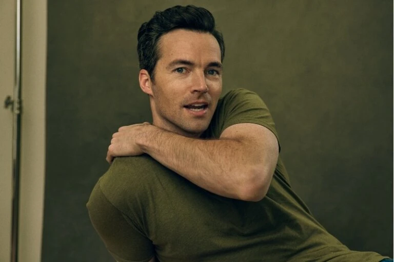 Portrait of a casually dressed adult male with dark hair, looking towards the camera against a muted olive-green studio backdrop.