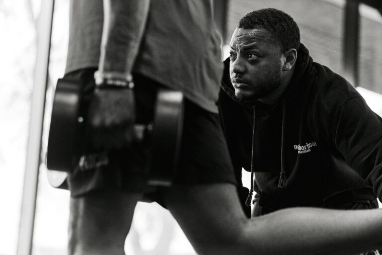 Monochrome shot of a man lifting a dumbbell with another man in a black hoodie observing during an indoor workout session.