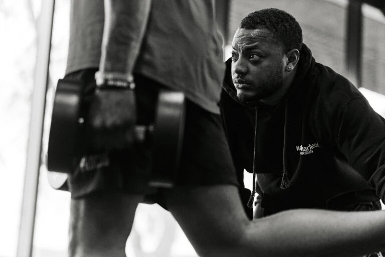 Monochrome shot of a man lifting a dumbbell with another man in a black hoodie observing during an indoor workout session.