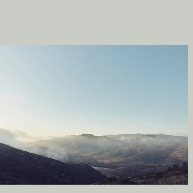 Mountain landscape featuring rolling hills partially obscured by low-lying fog under a clear blue sky, creating a serene and atmospheric vista.