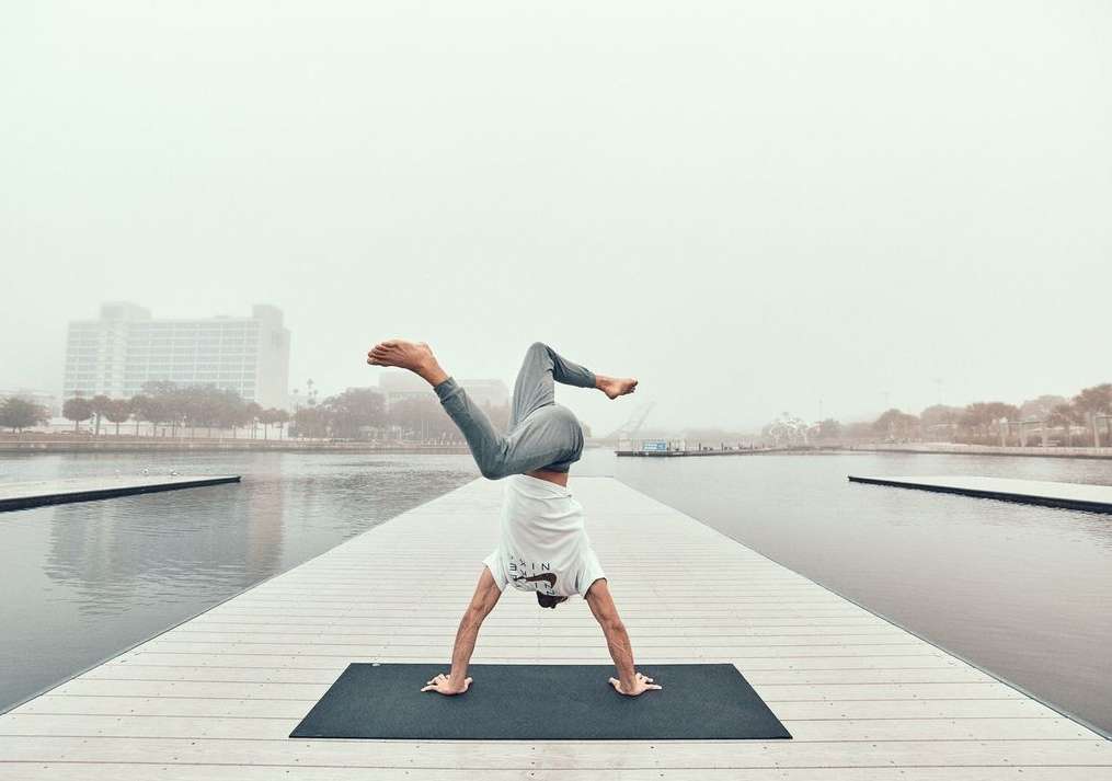 Adult man doing yoga handstand on a mat on a dock, wearing a white shirt and grey pants with a city and fog in the background.