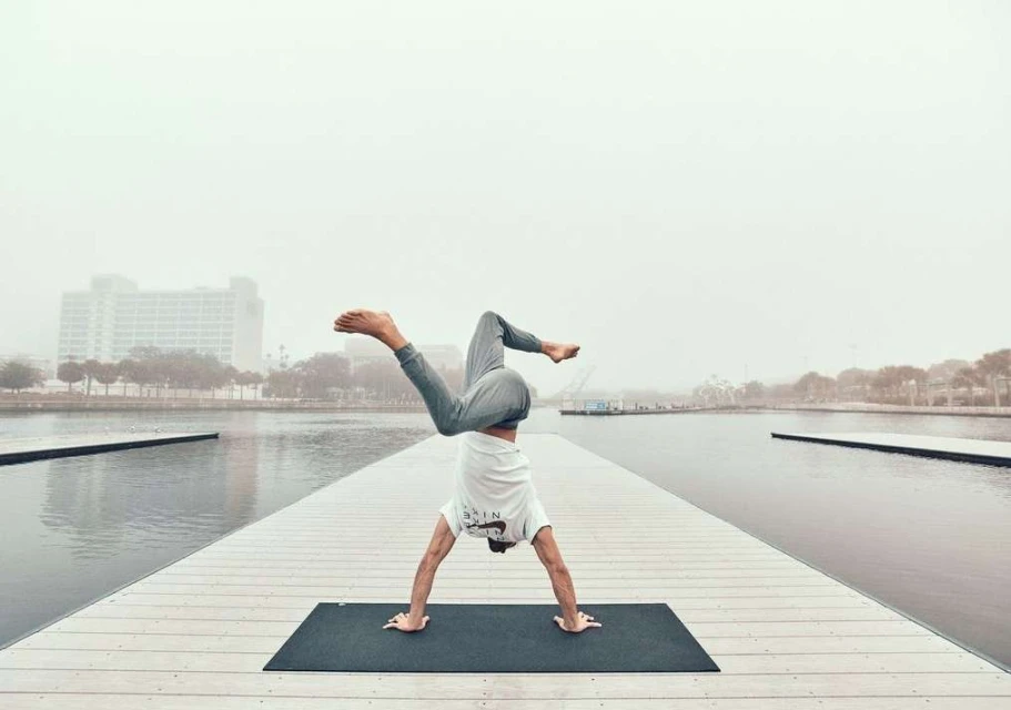 Adult man doing yoga handstand on a mat on a dock, wearing a white shirt and grey pants with a city and fog in the background.