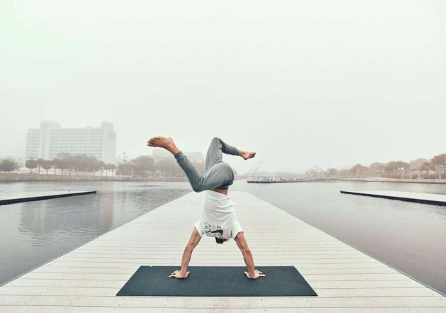 Adult man doing yoga handstand on a mat on a dock, wearing a white shirt and grey pants with a city and fog in the background.