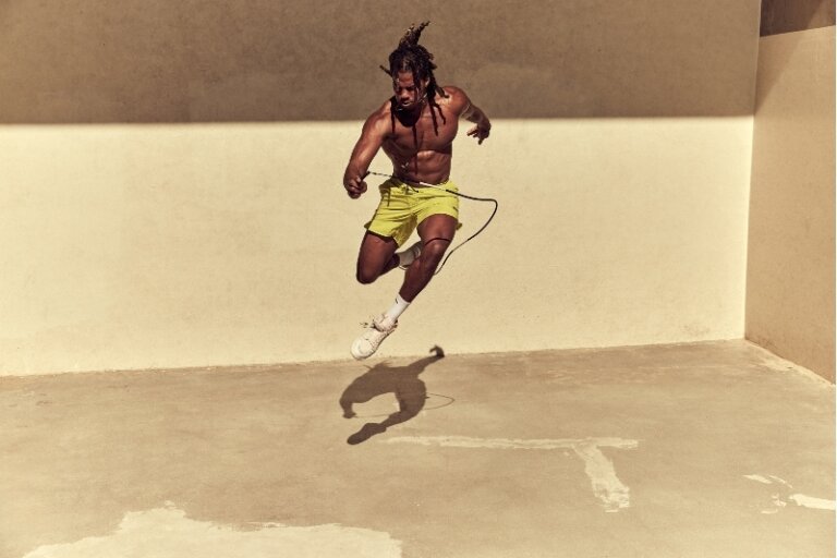 Athletic, shirtless man jumping rope in an indoor space with cream-colored walls and concrete floors, demonstrating fitness and athleticism.