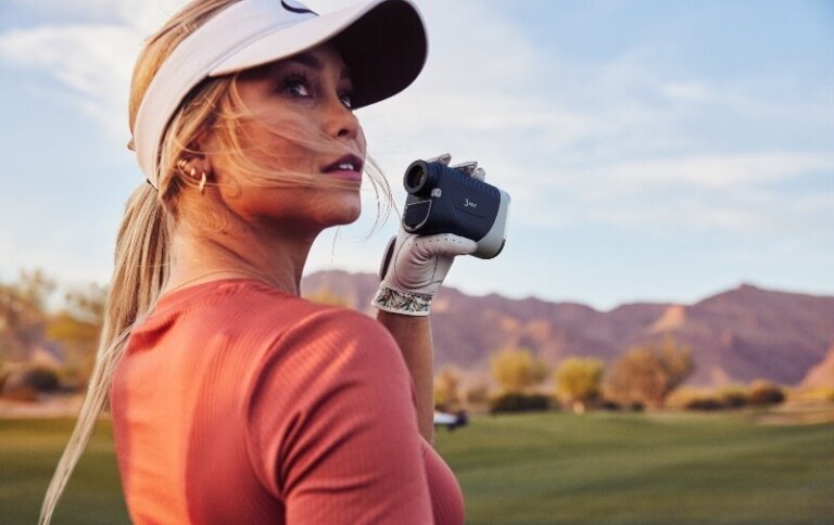 Woman in visor using rangefinder on golf course with mountains in the background on sunny day, wearing t-shirt and golf glove.