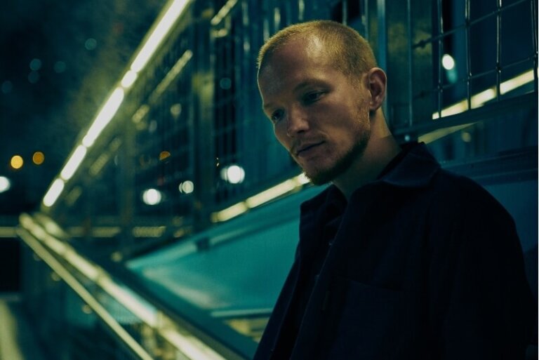 Adult man with a slight smile in a dark interior with turquoise metal structural elements and soft focus. Medium shot, portrait orientation.