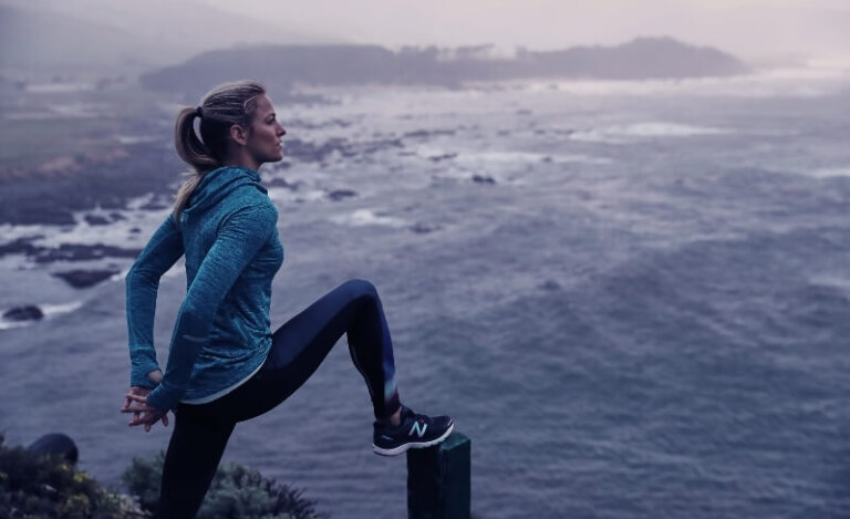Adult woman in athletic wear performs leg stretches on a stone ledge overlooking the ocean under a cloudy sky during daytime.