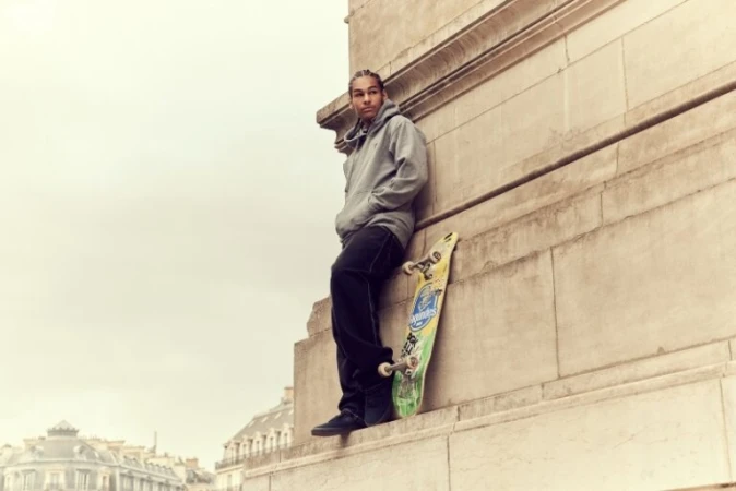 Young man in a grey hoodie leaning against a stone building ledge with a skateboard next to him