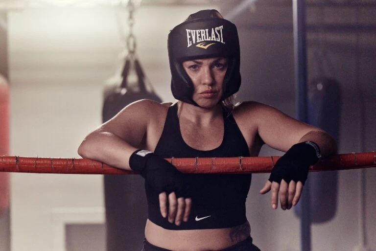 Woman boxer in Everlast headgear leans on ropes in boxing ring with punching bag in background; she wears black workout clothes and gloves.