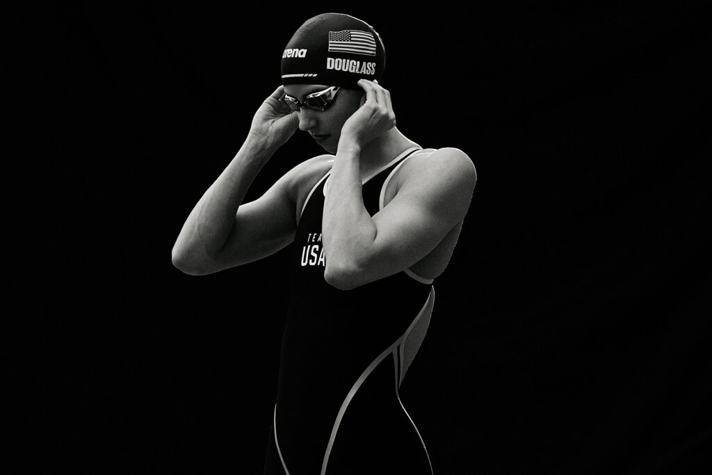 A female athlete in a USA swim cap and swimsuit, wearing goggles, adjusts her swim cap in a studio setting with dark background.