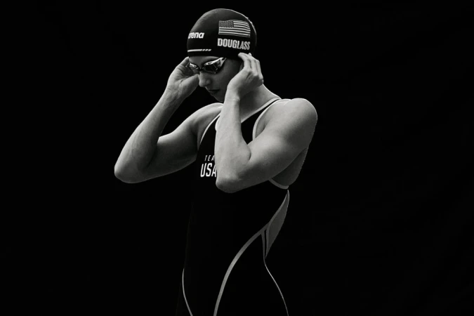 A female athlete in a USA swim cap and swimsuit, wearing goggles, adjusts her swim cap in a studio setting with dark background.