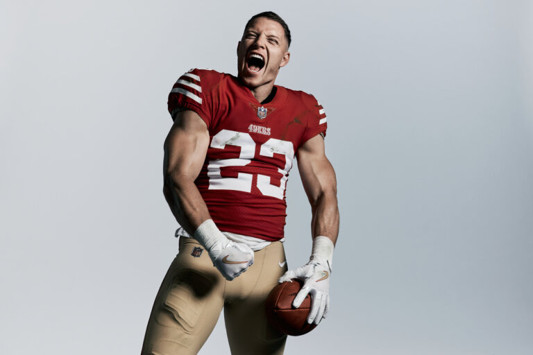 American football player in a red jersey shouting and holding a football against a grey background
