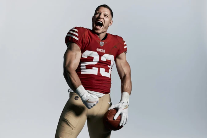 American football player in a red jersey shouting and holding a football against a grey background