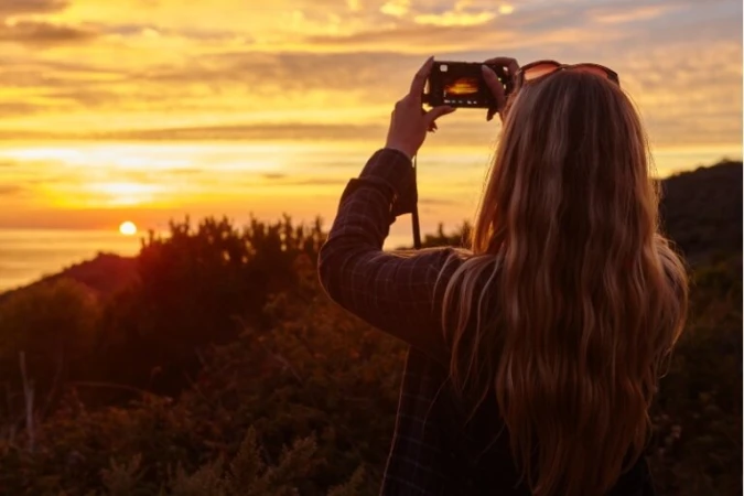 Woman with long blonde hair holding a camera up to photograph a vibrant sunset over the ocean, viewed from behind.