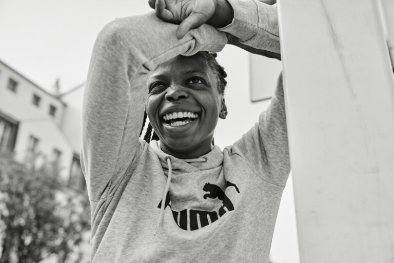 Smiling Black woman in a grey hoodie with arms raised and hands clasped behind her head, photographed by Coty Tarr.