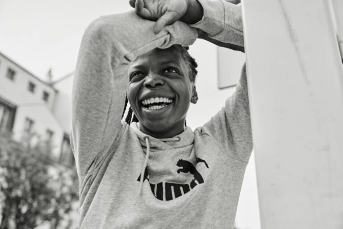 Smiling Black woman in a grey hoodie with arms raised and hands clasped behind her head, photographed by Coty Tarr.