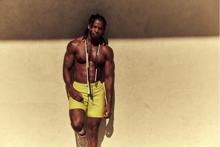 Athletic man in yellow shorts stands against wall with skipping rope around neck in a medium outdoor shot. Fitness and exercise theme.