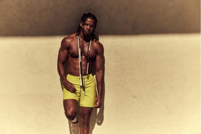Athletic man in yellow shorts stands against wall with skipping rope around neck in a medium outdoor shot. Fitness and exercise theme.