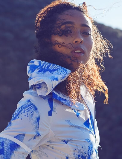 Portrait of a woman with curly hair wearing a blue and white patterned hooded jacket against an outdoor backdrop with muted tones.
