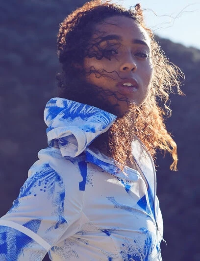 Portrait of a woman with curly hair wearing a blue and white patterned hooded jacket against an outdoor backdrop with muted tones.