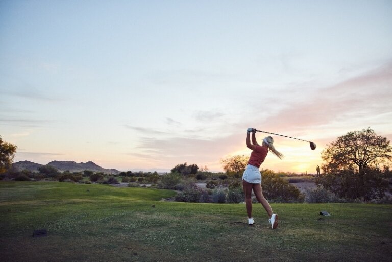 A woman in a red shirt and white skirt swings a golf club on a sunny golf course with trees and mountains in the background.