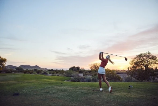 A woman in a red shirt and white skirt swings a golf club on a sunny golf course with trees and mountains in the background.