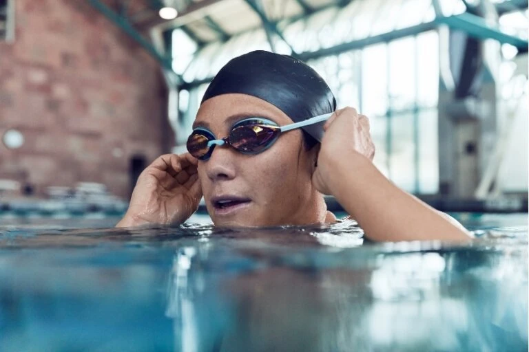 A woman with a swim cap and goggles standing in the shallow end of a swimming pool, with brick architecture visible in the background.