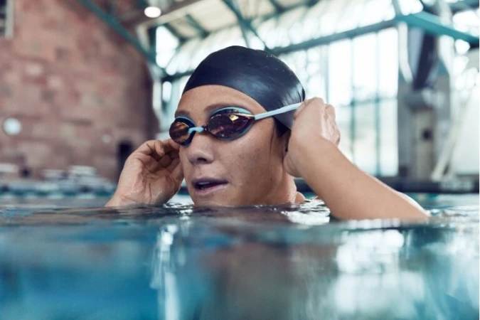 A woman with a swim cap and goggles standing in the shallow end of a swimming pool, with brick architecture visible in the background.