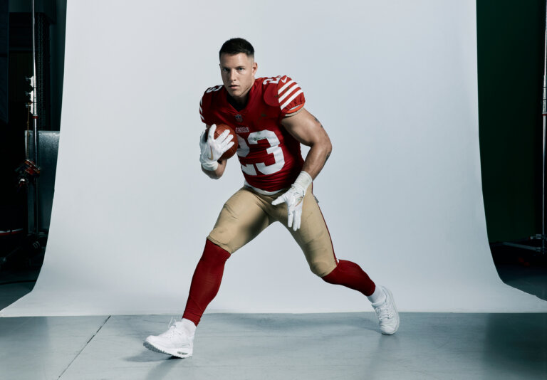 An American football player in uniform, wearing white gloves and shoes, is running and holding a football in a studio setting.
