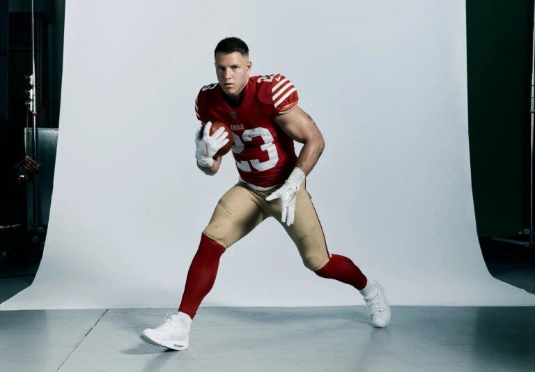 An American football player in uniform, wearing white gloves and shoes, is running and holding a football in a studio setting.