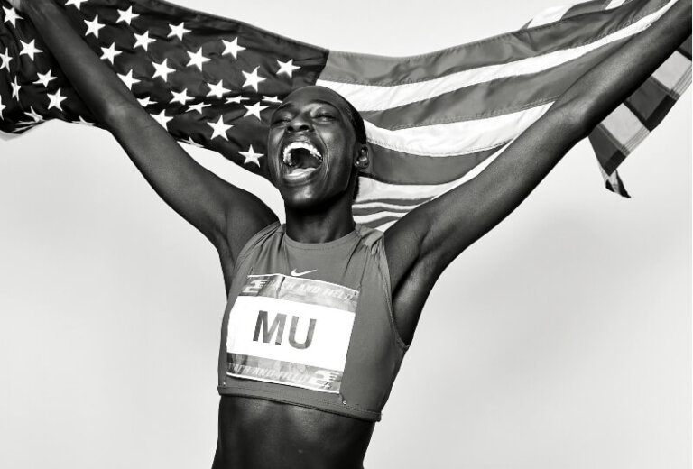 Black and white portrait of a female athlete holding an American flag and celebrating a victory, wearing a sports uniform.