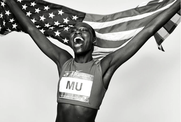 Black and white portrait of a female athlete holding an American flag and celebrating a victory, wearing a sports uniform.