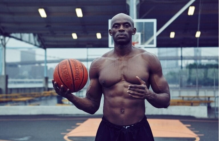 Shirtless adult male athlete holds a basketball on an outdoor court with city buildings in the distance during the daytime.
