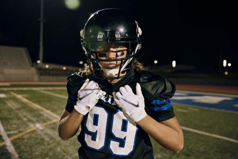 Football player in uniform and helmet stands on a field at night under bright stadium lights, holding their white-gloved hands up.