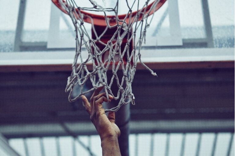 Close-up of a hand reaching up to a basketball net with an orange rim, set against a blurred urban outdoor background.
