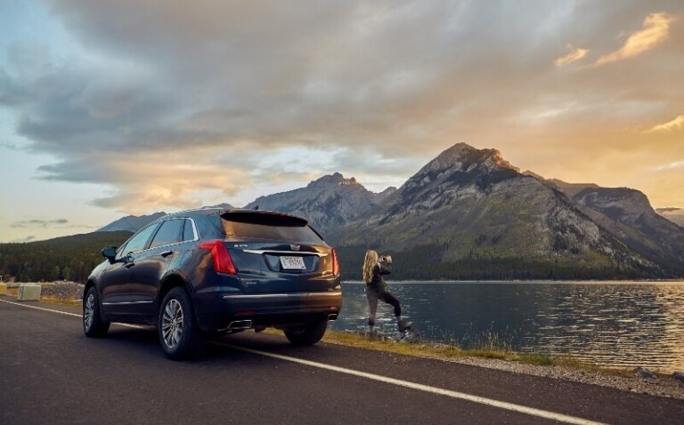 Dark-blue SUV parks on a road with a lake and mountains in the background. A woman takes a photo of the scenery with a camera.