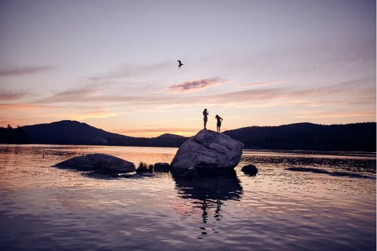 Two silhouetted figures stand atop a rock formation surrounded by water at dusk with a bird flying overhead, silhouetted mountain range.