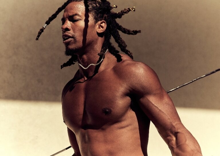 Toned and muscular Black man with dreadlocks gazes off-camera, wearing a delicate necklace against a neutral background, holding a spear.